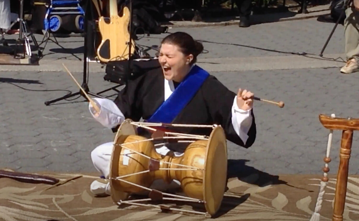 Melanie Ehrlich playing janggoo, an hourglass-shaped traditional Korean drum, with eyes shut and mouth open in a smiling yell of excitement
